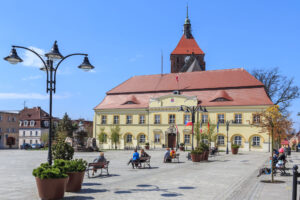 Rynek z zabytkowym ratuszem i wieżą kościoła w tle, fontanną, ławkami i donicami z zielenią